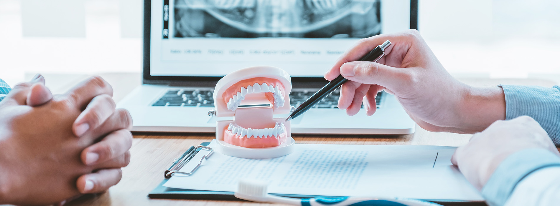 A person holding a tooth model while another examines it, with a laptop displaying medical images in the background.