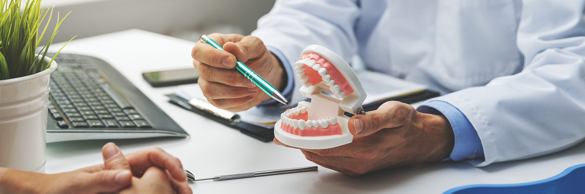 The image shows a dental professional holding a model of a human mouth, with another person looking at it and a third person writing on a clipboard. They are in an office setting with a desk, keyboard, and plants visible in the background.