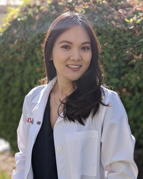 The image is a photograph of a person standing outdoors, smiling and posing for the camera. They are wearing a white lab coat over a dark top and have short hair.