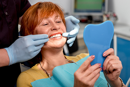 A woman is seated in a dental chair, smiling and showing off her teeth, while holding a blue model tooth with a dentist standing behind her, both wearing face masks.