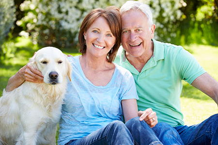 A man and woman are sitting outdoors, smiling and posing with a golden retriever between them.