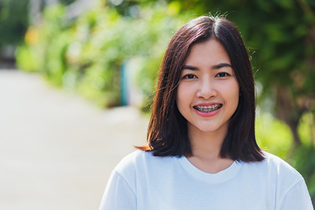 The image shows a woman with short hair, smiling at the camera. She is wearing a white t-shirt and has her hair styled in a bob cut. The background features a blurred outdoor setting with greenery.